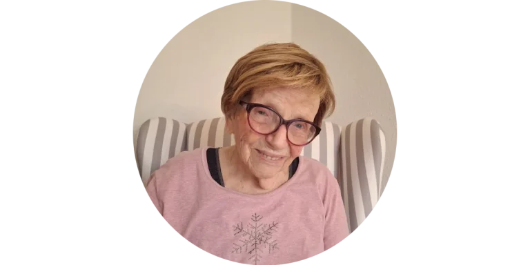 Lächelnde Kundin von Mia Oppermanns Seniorenassistenz Lächelnde ältere Dame mit Brille und oranger Strickjacke sitzt entspannt auf dem Sofa – Kundin von Mia Oppermann.