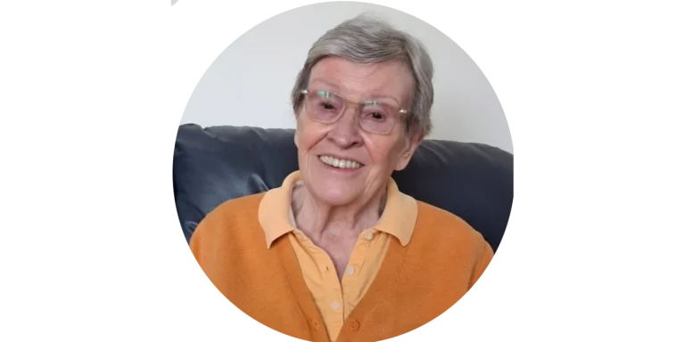 Lächelnde Kundin von Mia Oppermanns Seniorenassistenz Lächelnde ältere Dame mit Brille und oranger Strickjacke sitzt entspannt auf dem Sofa – Kundin von Mia Oppermann.